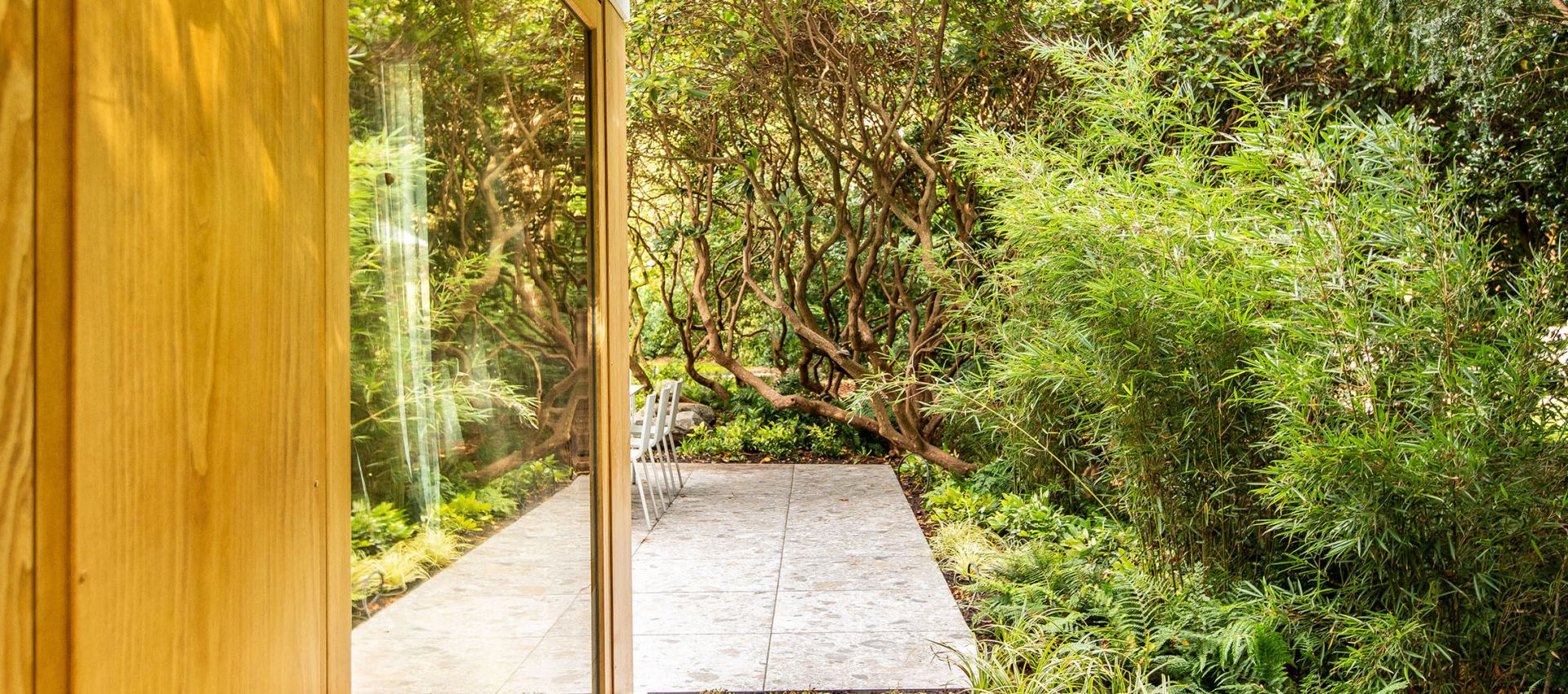 Holzhaus und große Fensterfront mit Blick in Richtung eines Sitzplatzes mit üppger Bepflanzung im Waldcharakter